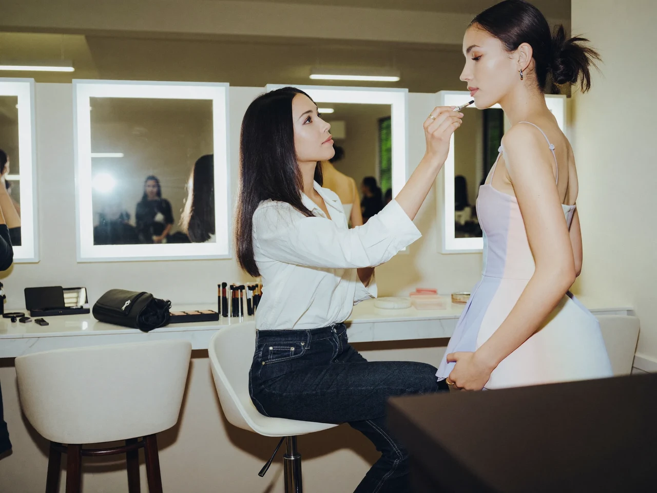 Lyst makeupstudie med flere arbejdsstationer, spejle og en elev, der øver på en model